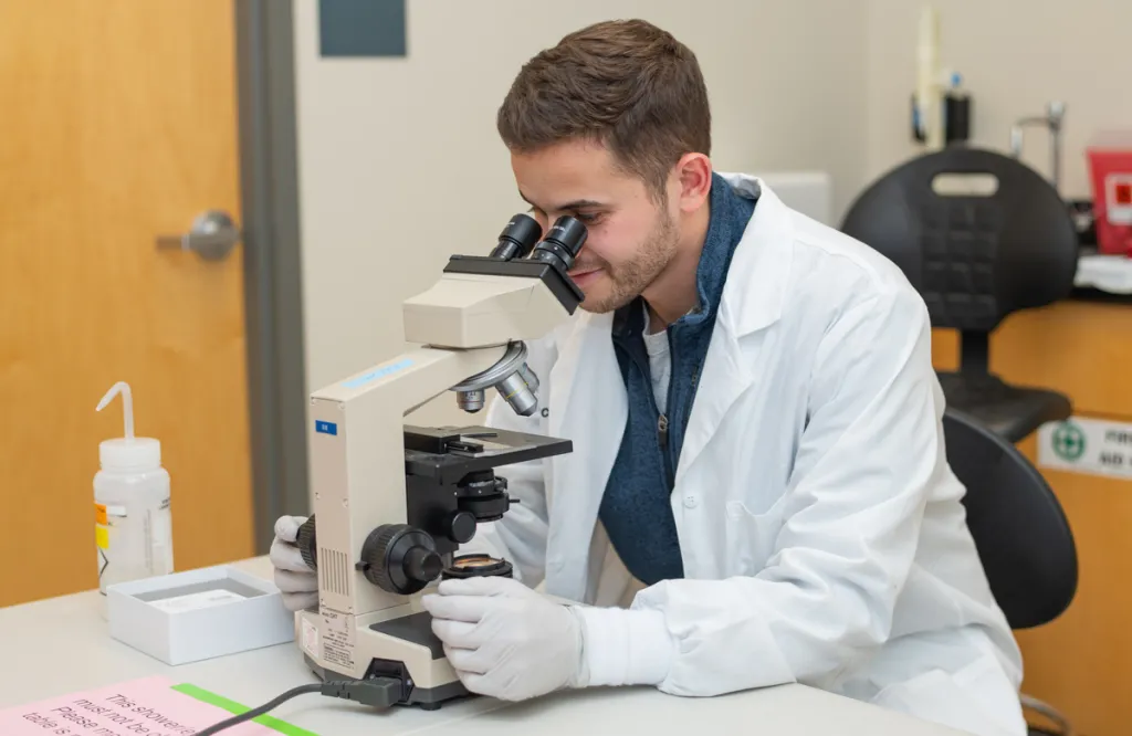 A student in a white lab coat peers into a microscope