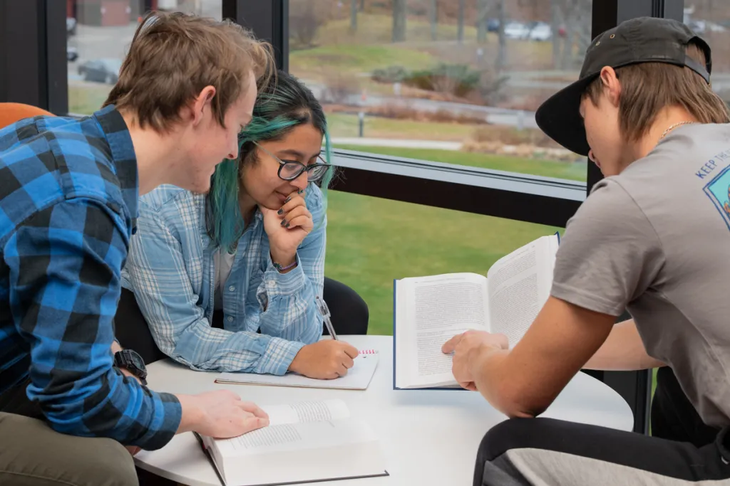Three students sit around a table with open books scattered across the top