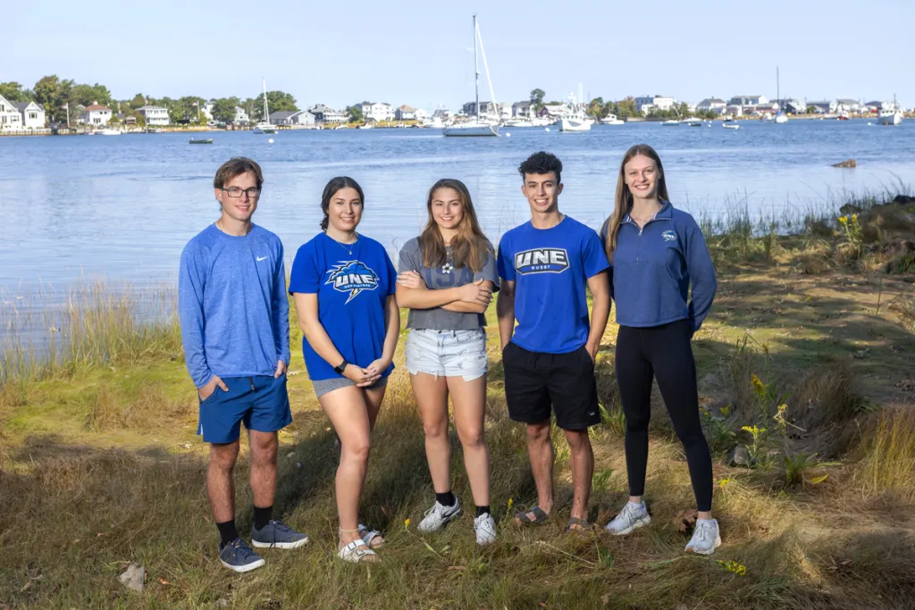 Five students pose together in front of a river with a small Maine town on the other side