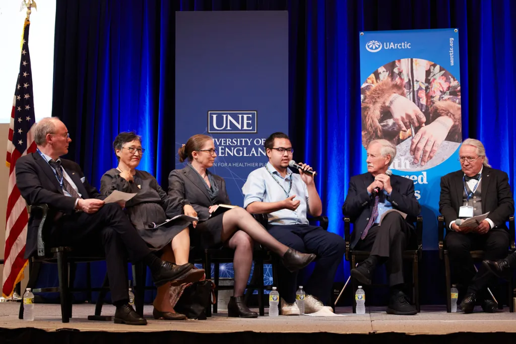 A man speaks at a U Arctic event alongside five other speakers on stage, including United States Senator of Maine, Angus King