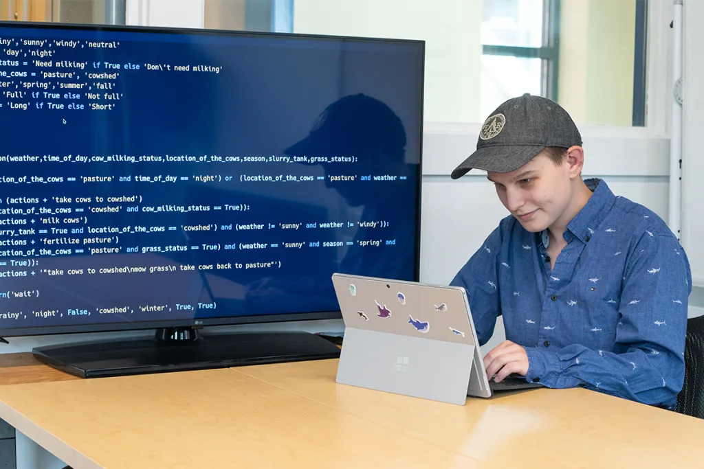 A student works on code on their laptop next to a large monitor displaying their work live