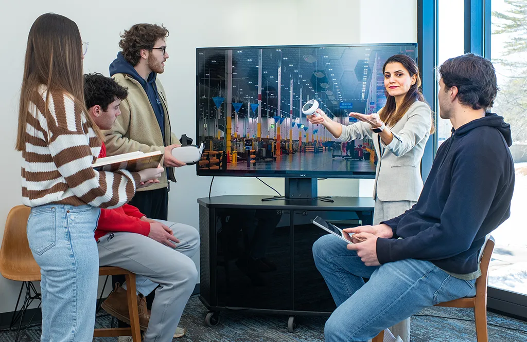 A group of four students listen intently to their professor as she describes a virtual warehouse image on a large monitor