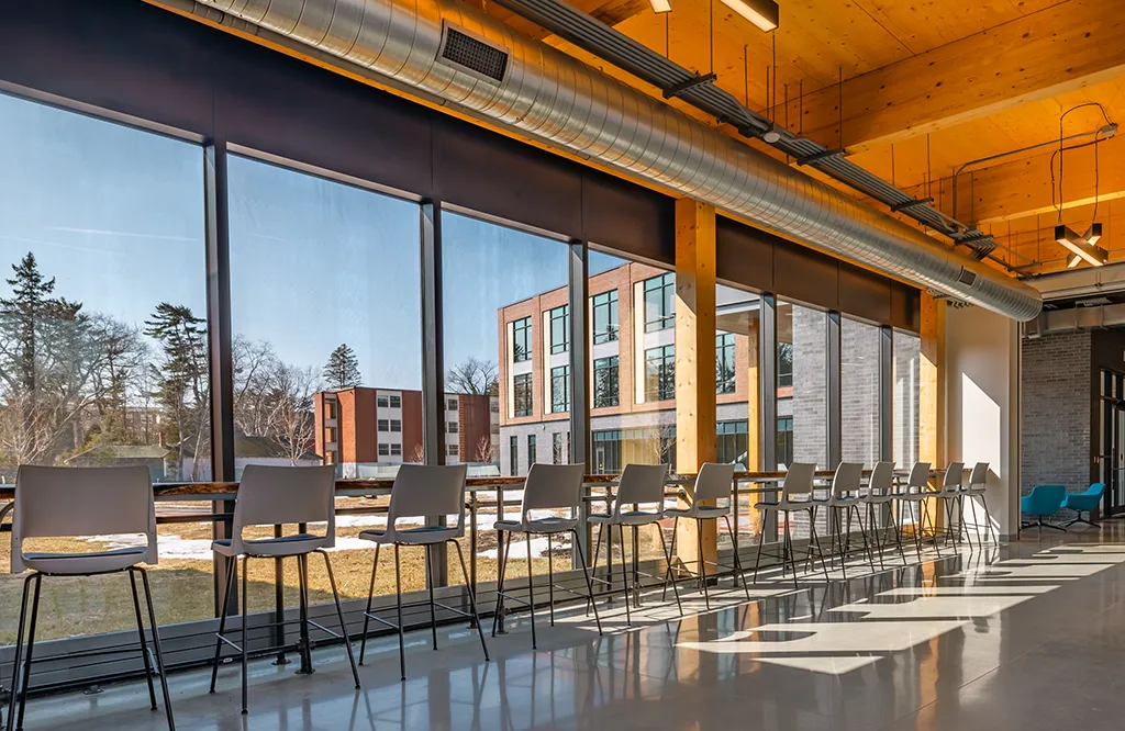 Several high-top chairs lined up at a wooden counter in front of a row of large windows looking out at a green quad