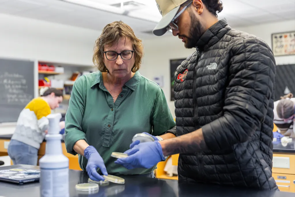 A student shows their petri dish of yeast bacterial colonies to their professor during lab
