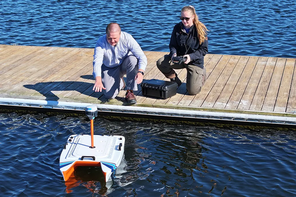 A student and their marine science professor drive a remote water sampling boat from a dock