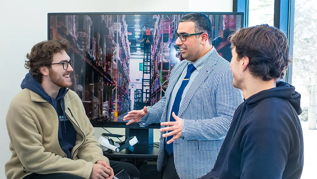 A professor speaks with two C O B students in front of a t.v. monitor displaying a virtual warehouse