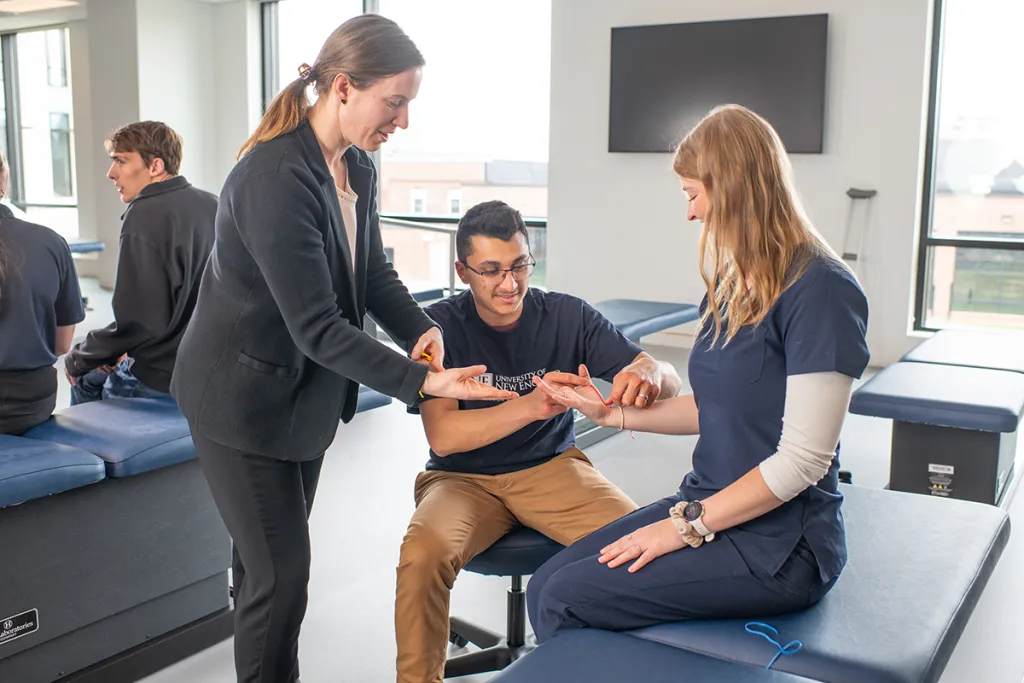 An O T professor assists a student as he uses a rubber band to learn about muscle activation and movement on another student's wrist