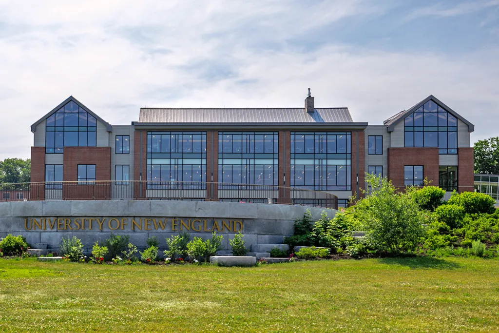 A brick building with numerous windows sits behind a stone sign that reads, "University of New England"