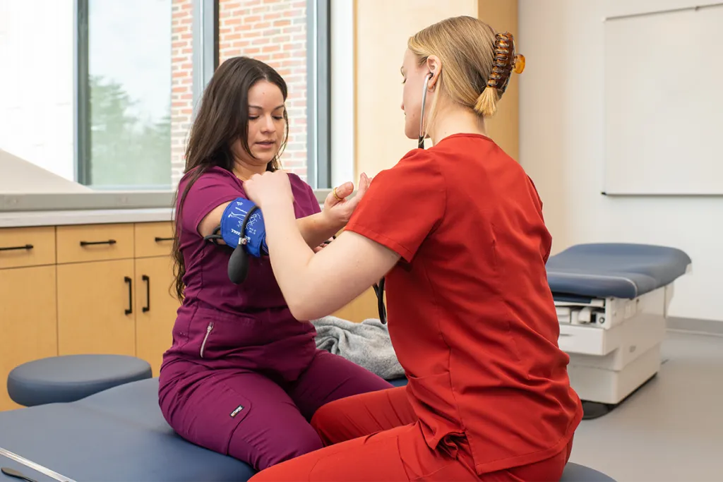 A C O M student practices getting a patient's blood pressure on another C O M student in a clinical skills lab setting