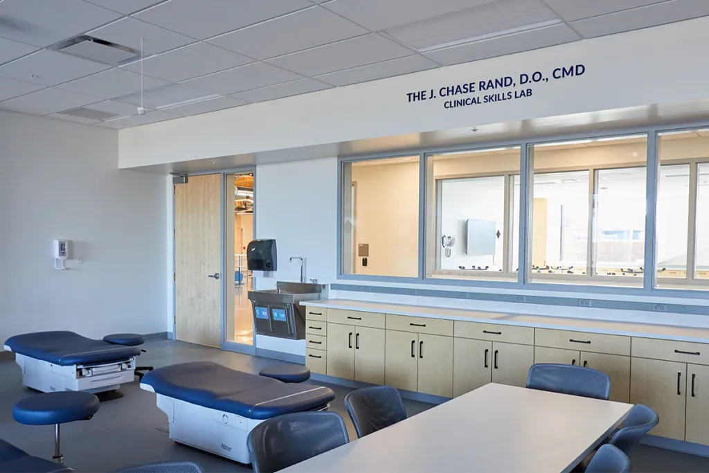 Two blue examination tables and white rectangular table in front of a row of light-wood cabinetry and large windows facing into a hallway