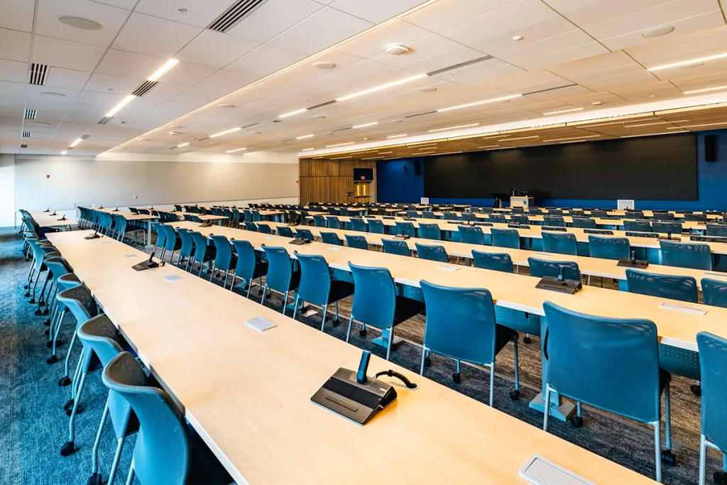 A large monitor extending most of a wall sits behind several rows of light-wood tables and blue chairs