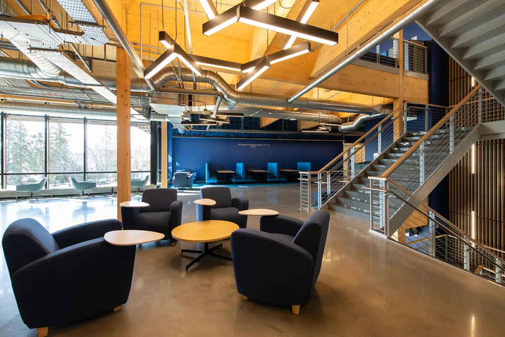 Modern office lounge with dark blue chairs, small round tables, large windows, exposed ducts, and a metal staircase against a blue accent wall