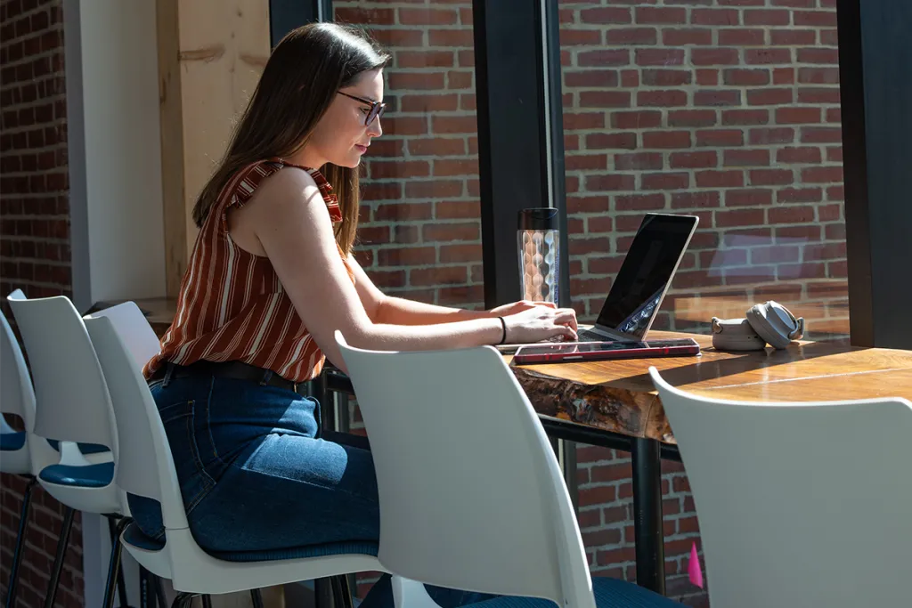 A student studies on her laptop at a long wooden table in front of large windows overlooking a brick building
