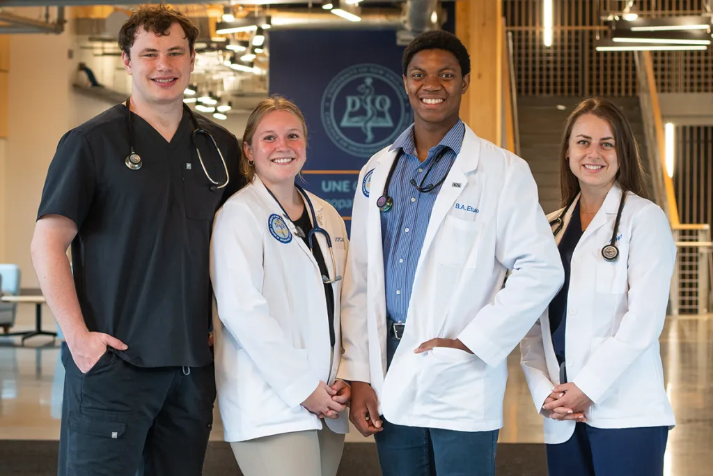 A group of C O M students, one in scrubs and three wearing white coats, pose in front of a hallway with cement floors and industrial ceilings