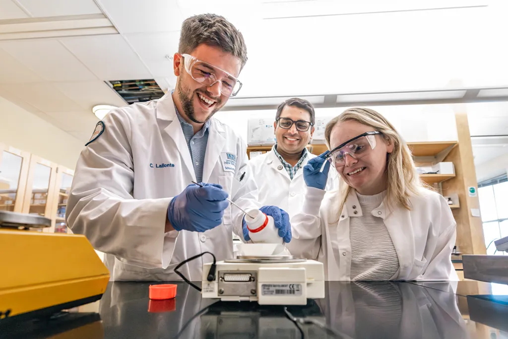 Two students work with a professor in a lab, all wearing white coats and gloves as they measure powder on a scale