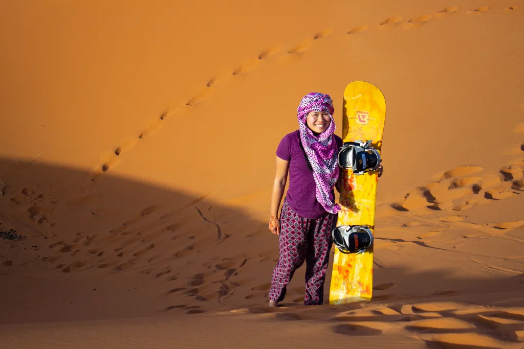 A student holds a sand board while standing in a desert