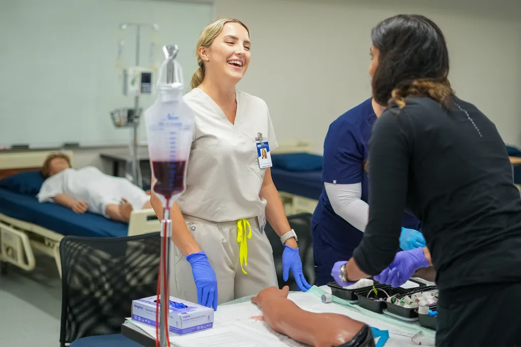 A C O M student in scrubs preps a patient simulator arm for an I V