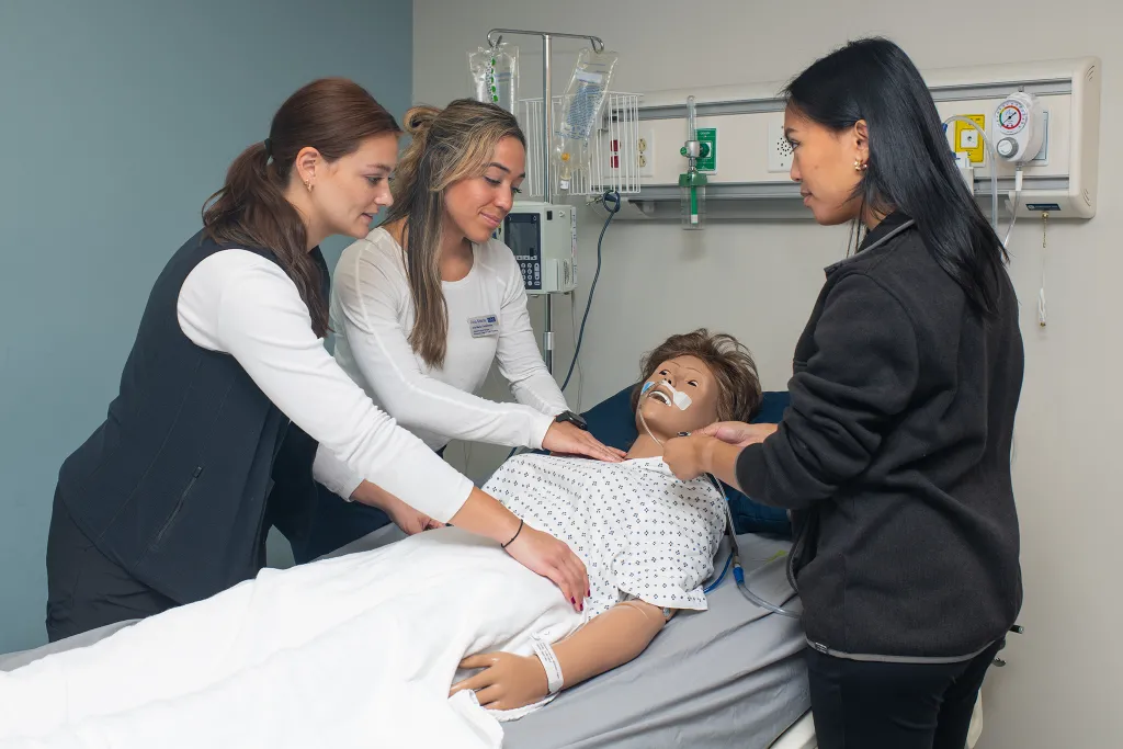 Three U N E healthcare students collaborate around a medical training mannequin in a hospital simulation room. The mannequin patient lies on a hospital bed with IV equipment and medical monitoring devices visible in the background, demonstrating hands-on interprofessional clinical training.