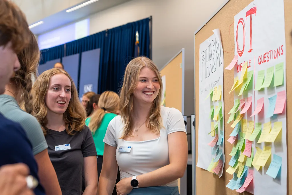 Two University of New England students wearing name tags stand next to an interactive poster board covered with colorful sticky notes organized under "OT" and "Nursing Questions" sections.