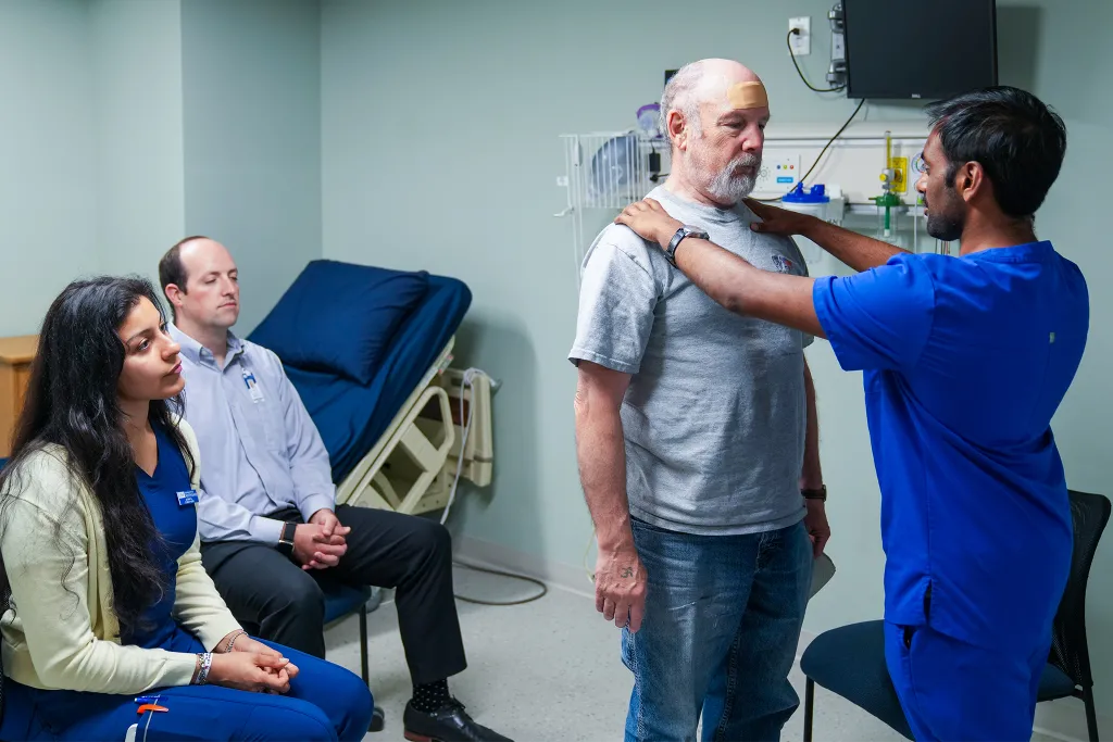 In a clinical training room, a U N E healthcare student in blue scrubs performs a shoulder examination on an older male patient in a gray t-shirt, while two other students observe from chairs nearby. Medical equipment and an examination table are visible in the background of this interprofessional learning scenario.