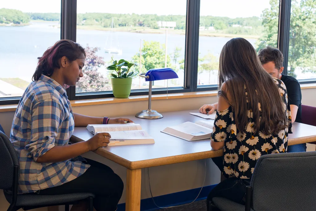 Three U N E students studying at a table next to large windows overlooking a river
