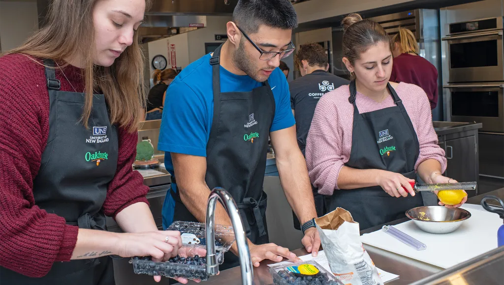 Three students in black aprons prep and wash various fruits and berries in the U N E class kitchen