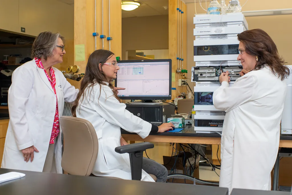 Houseknecht, Barlow, and a student researcher collaborate around a computer displaying data, with laboratory equipment visible in the background.