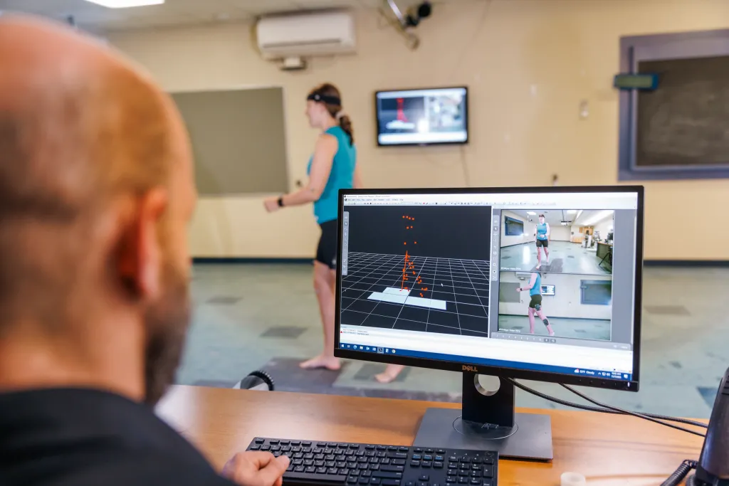 A professor analyzes motion capture data on a computer monitor showing 3 D biomechanical tracking points while a participant performs movement in the background