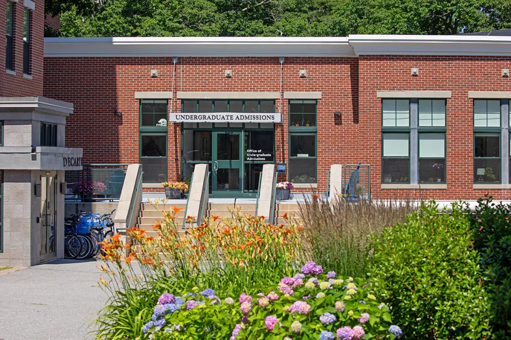 Brick building with "Undergraduate Admissions" sign above glass entrance doors, surrounded by colorful flowering plants and landscaping on campus.