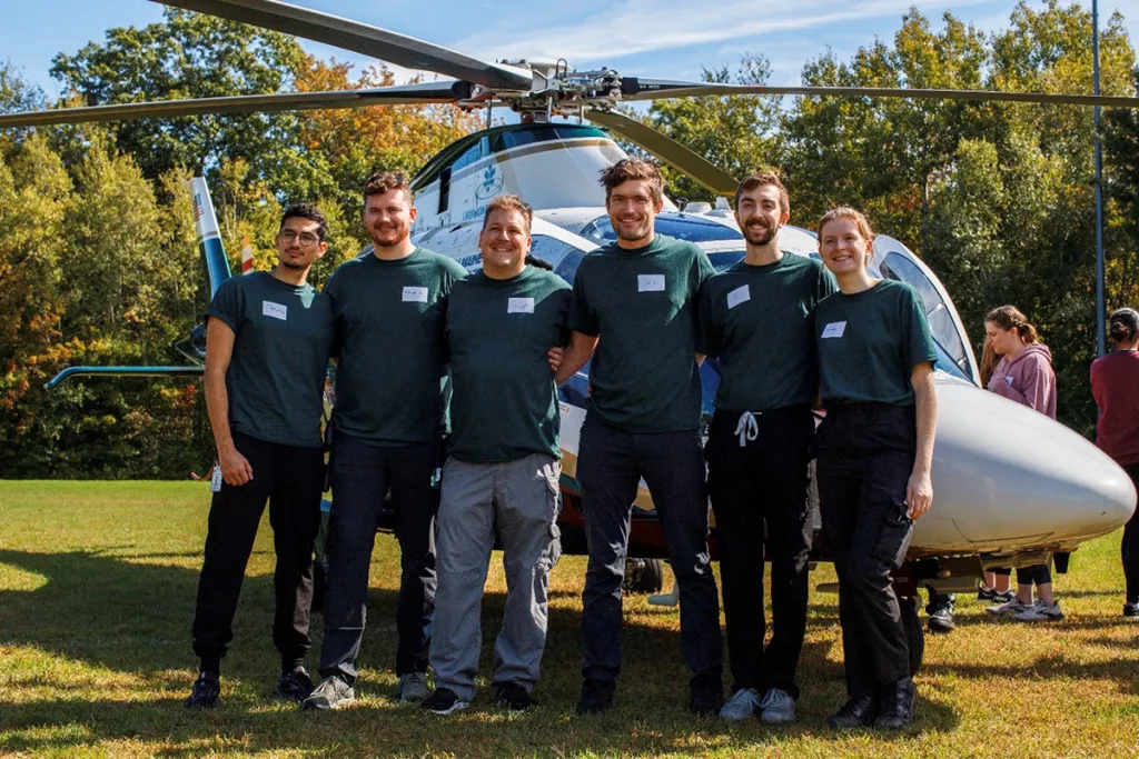 Six people in matching green shirts pose in front of a medical helicopter on a grassy field.