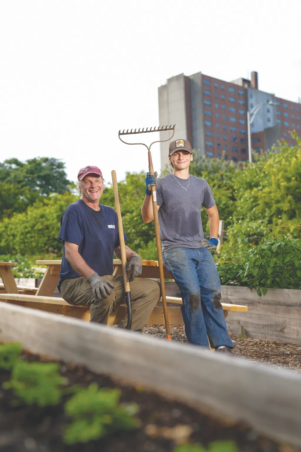 Luke Jenkins (Biology, ’26) and Cameron Wake, Ph.D. pose with gardening tools at a community garden in Portland, Maine, with campus buildings visible in the background.