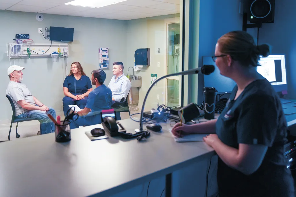 An ISIC team member monitors an interprofessional case with Michael, a patient actor, through an observation window in a clinical simulation laboratory.