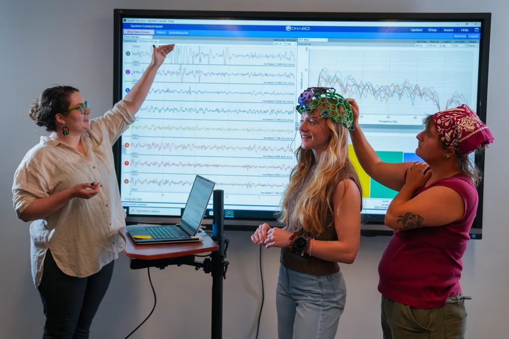 Sophia Crockett-Current points to brainwave data on monitor while Milo Lypps fits Jessica Howard with multicolored EEG sensor cap.