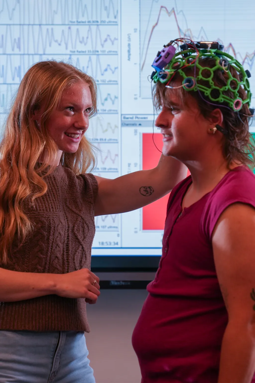 Jessica Howard fixes a colorful skull cap on Milo Lypps while standing in front of a monitor showing brain activity graphs