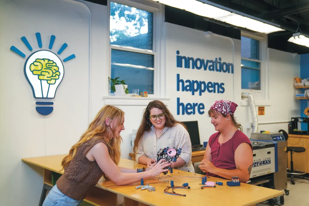 Jessica Howard, Sophia Crockett-Current, and Milo Lypps collaborate on projects at a table beneath "innovation happens here" signage in a makerspace.