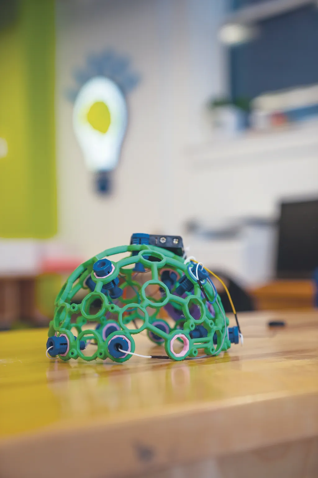 A green skull cap with electronic sensors sits on a table in a makerspace with a lightbulb logo visible in the background.