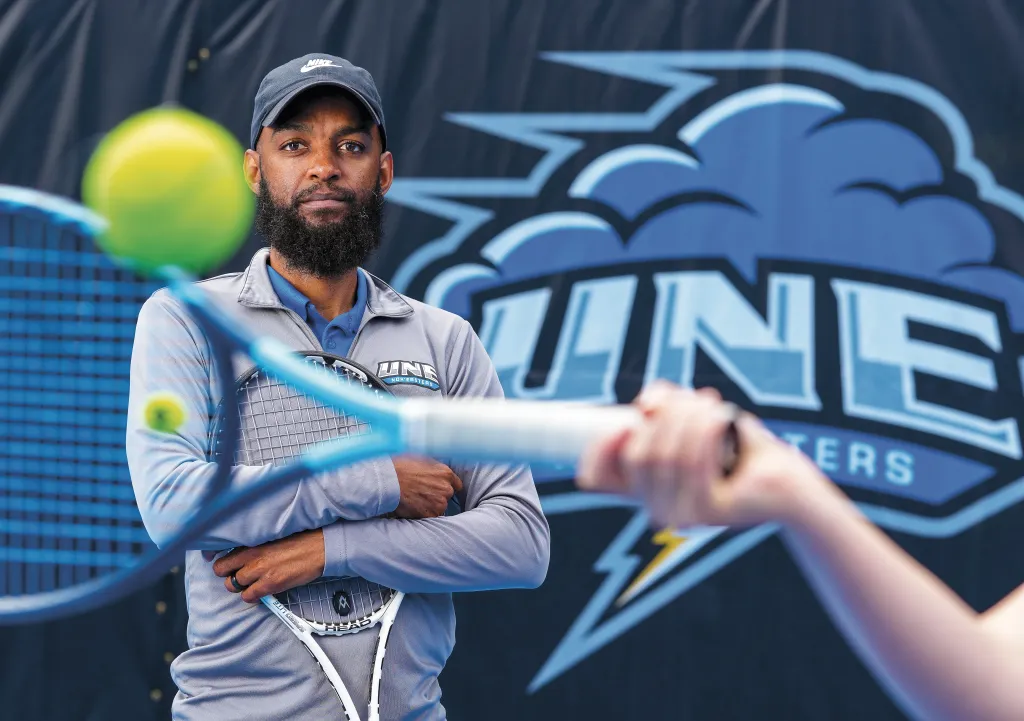 U N E men's and women's tennis head coach Jovan Jordan-Whitter holds a tennis racket in front of the U N E Nor'easters logo at the Blue Cyclone Complex.