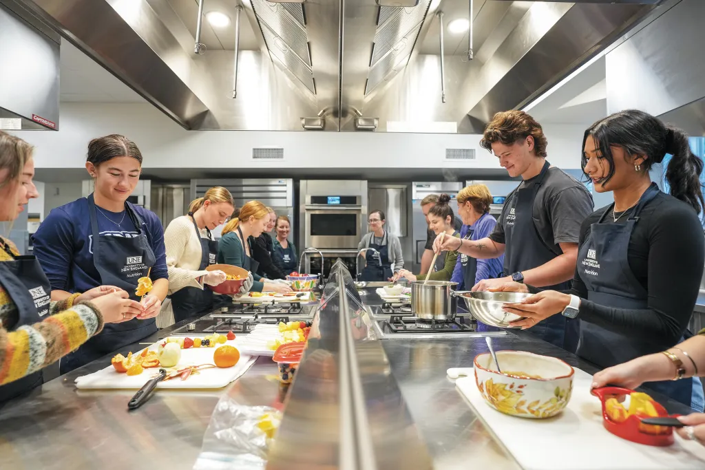 Students in aprons preparing food at stainless steel island in professional teaching kitchen with commercial appliances.