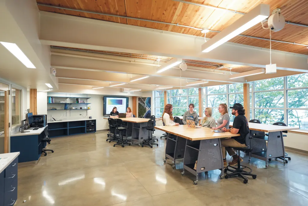 Collaborative learning space with wooden ceiling, floor-to-ceiling windows, students at tables, and mounted monitor displaying presentation.