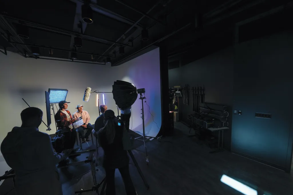 Film production crew with lighting equipment and cameras recording interview subjects against backdrop in studio.