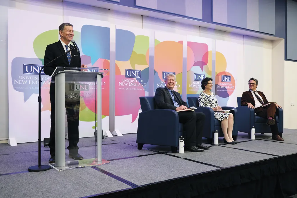 U N E President James Herbert speaks at a podium during the President's Forum while three panelists are seated on stage against a backdrop featuring colorful speech bubble graphics and U N E branding
