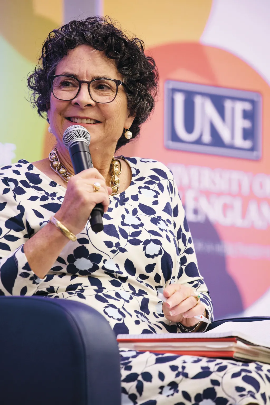 Jeanne A.K. Hey, moderator and U N E dean emerita, speaks into a microphone while seated during the President's Forum, wearing a black and white floral print dress