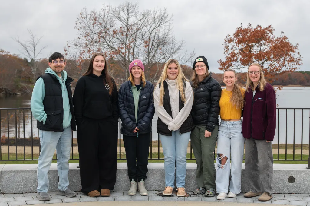 Seven students posing together on a campus overlook with autumn foliage and a lake visible in the background under an overcast sky.