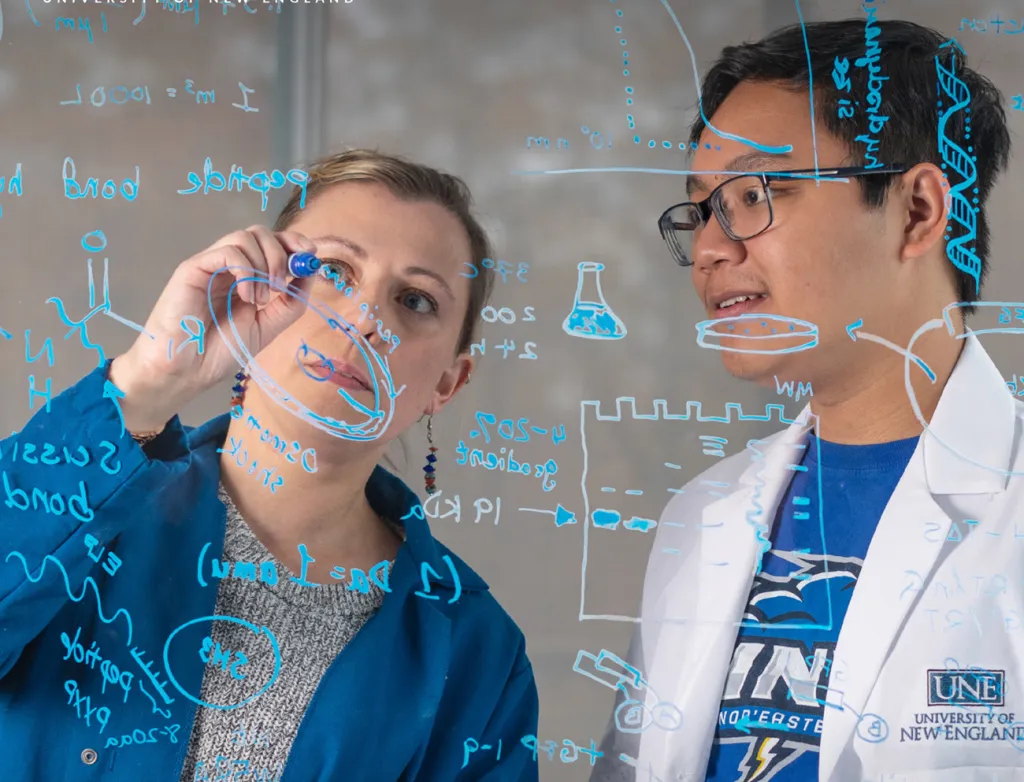 A professor in a blue jacket and student researcher in white lab coat examining scientific formulas and molecular diagrams drawn on transparent glass surface.