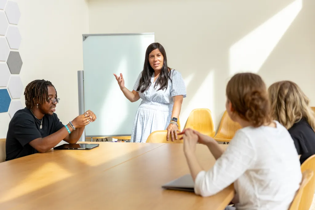 Professor Vlachos leading a small group discussion with students seated around a table in a bright, modern classroom with yellow chairs and a whiteboard.