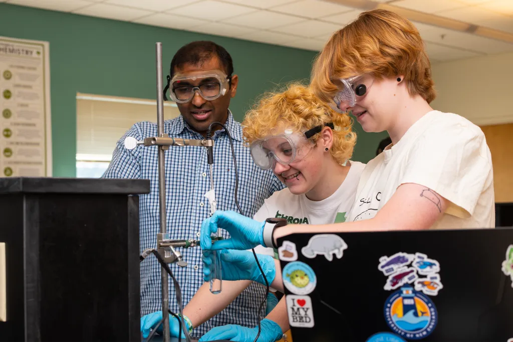 Chemistry professor guiding two students wearing safety goggles and blue gloves as they conduct a laboratory experiment with mounted equipment in a classroom setting.