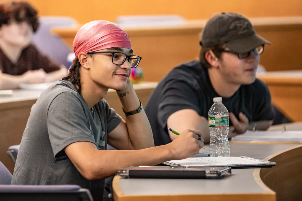 Students engaged in classroom discussion, with one student in a pink headwrap and glasses smiling while taking notes at their desk.