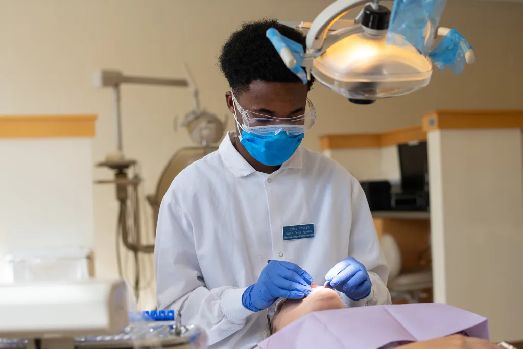 Dental hygiene student wearing protective mask, face shield, and blue gloves performing a teeth cleaning on a patient at U N E's Oral Health Center.