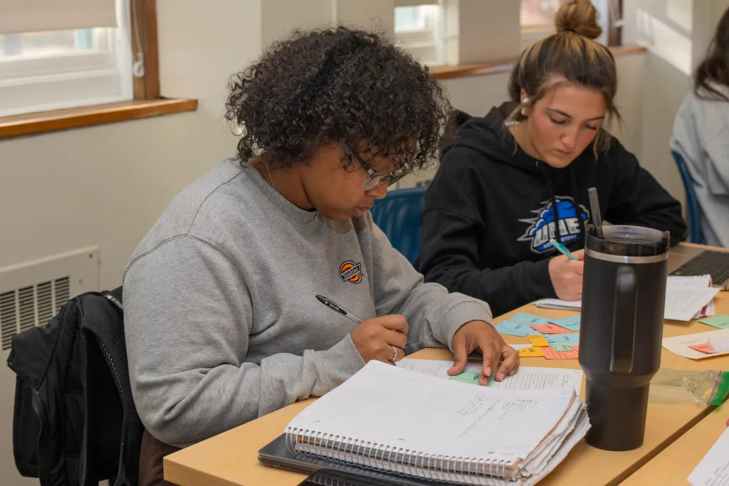 Students studying together at desks in a classroom, with notebooks, papers, and water bottles, focused on their coursework in a bright, naturally-lit space.