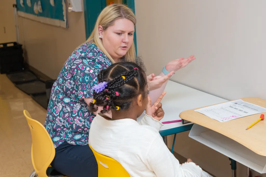 Education student helping an elementary student with math while seated at a yellow table in a classroom setting.